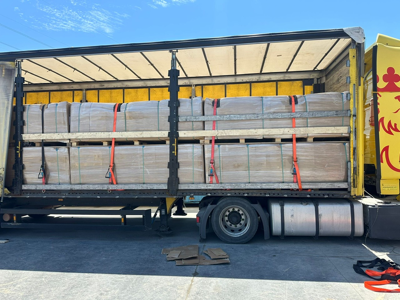 Yellow trailer loaded with palletized acoustic wall panels secured with orange ratchet straps
